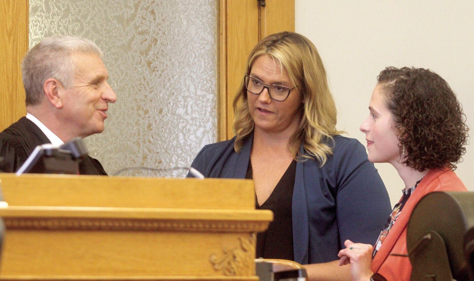 A judge leans from the bench to speak with two lawyers
