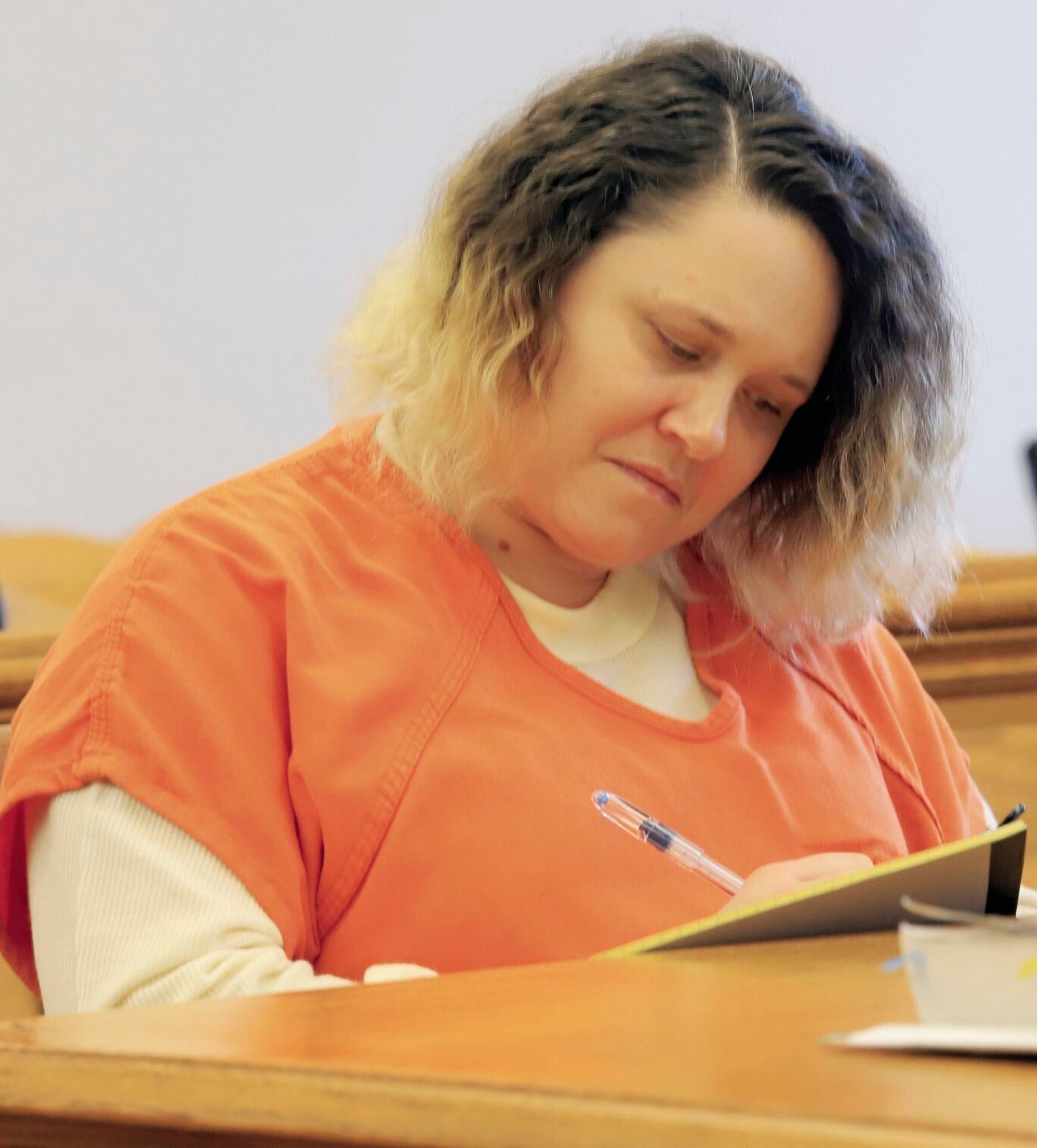 A woman in an orange jail uniform writes on a legal pad at a courtroom table